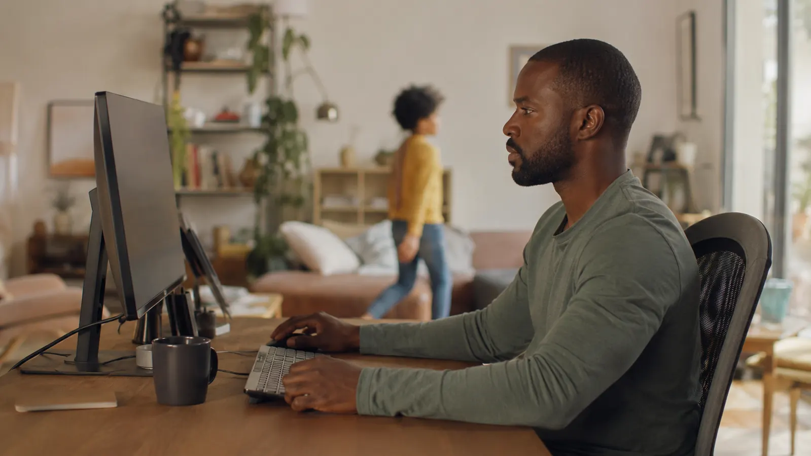 Person working at a home office desk with a child walking past behind them, screen not visible to the passerby