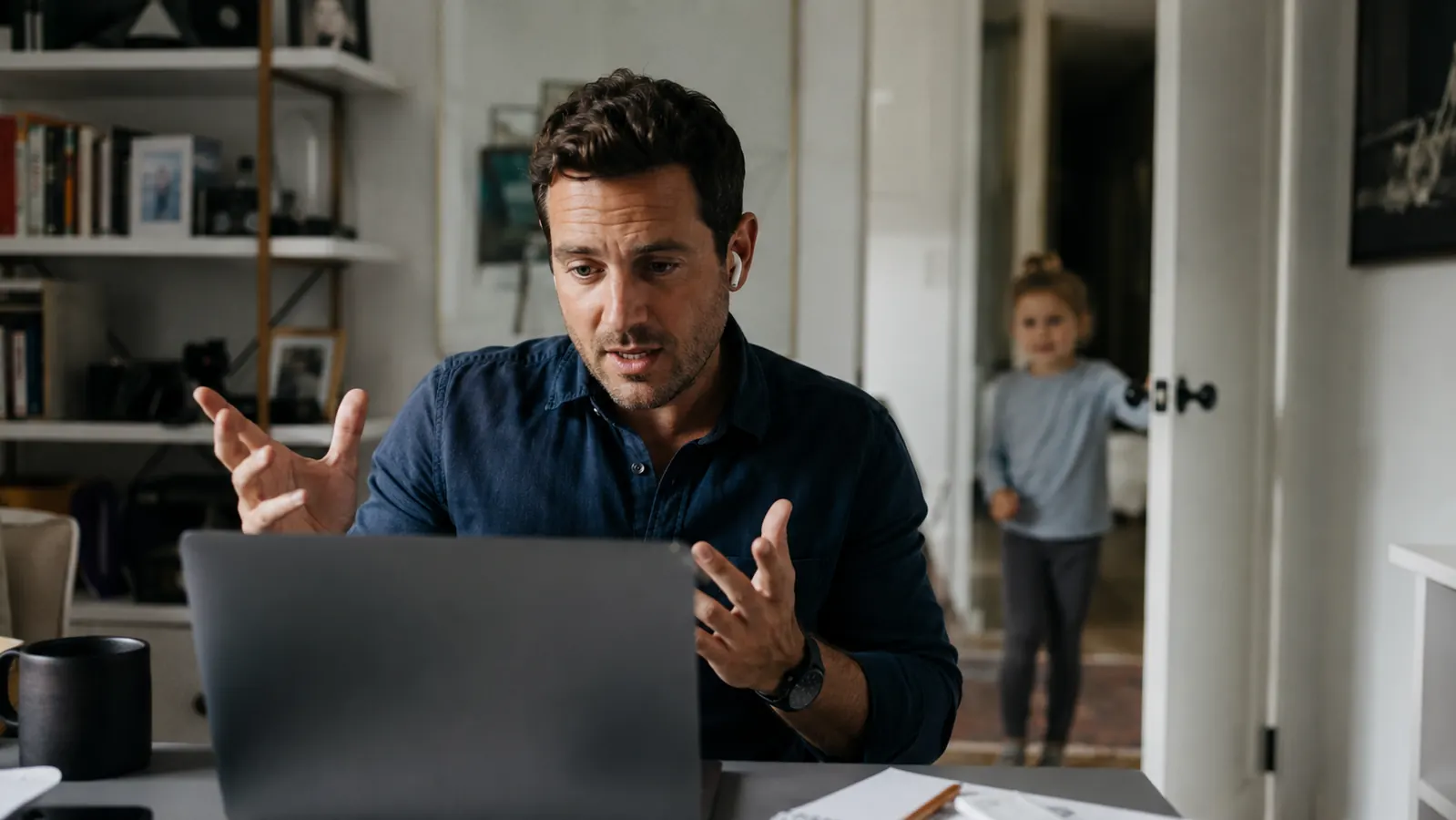 Father at a home office desk, young daughter peeking around the doorframe in the background, he is focused on a video call